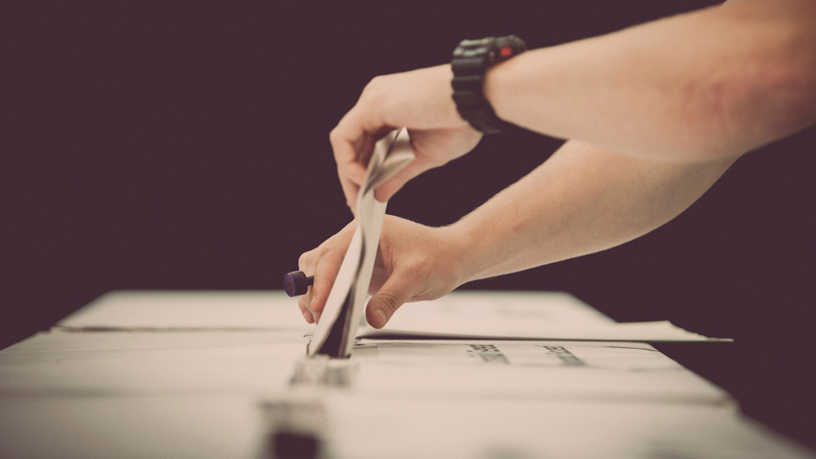 Jemand mit einer schwarzen Sportuhr am linken Handgelenkt steckt mit beiden Händen einen Stimmzettel in den Schlitz einer Wahlurne.