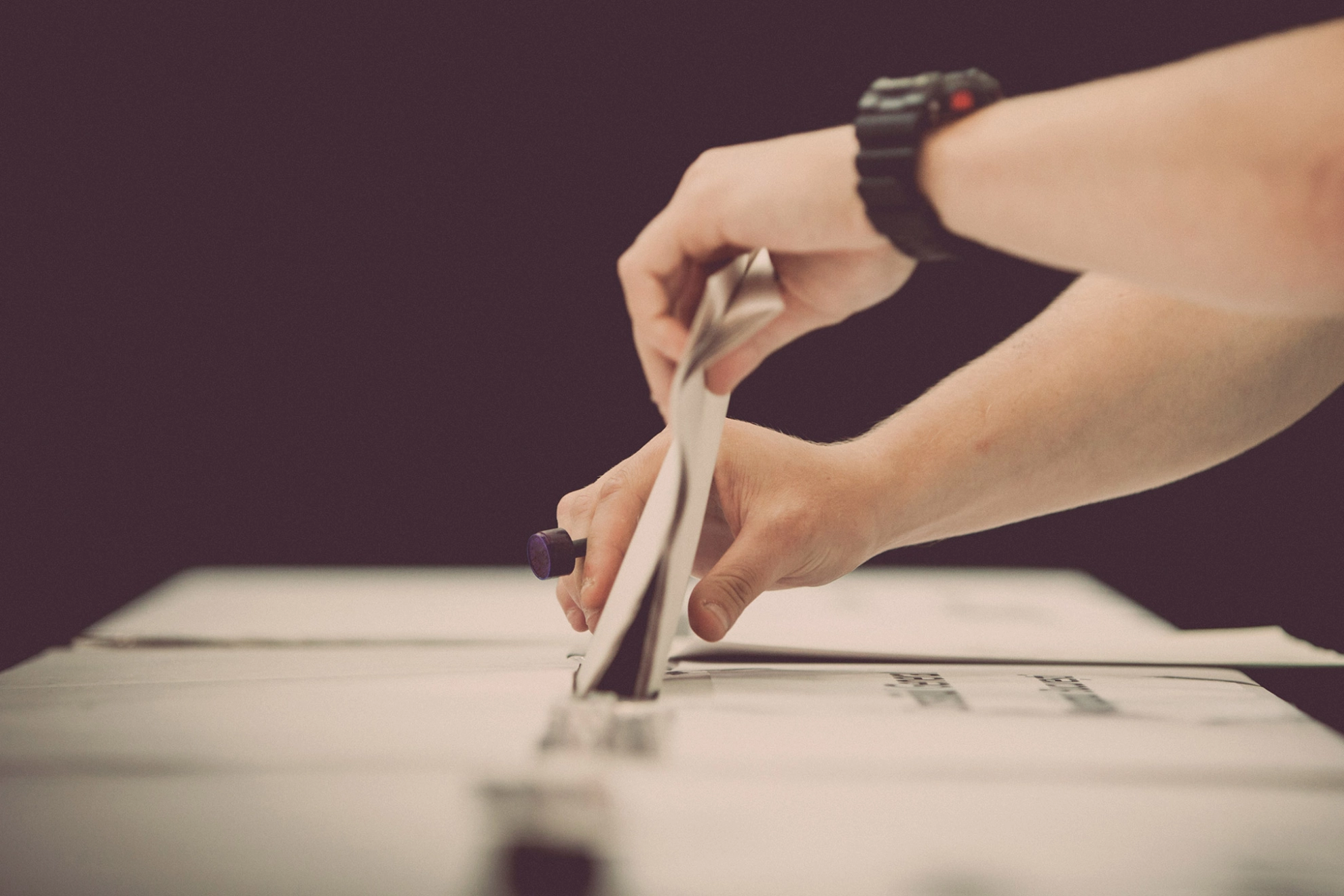 Jemand mit einer schwarzen Sportuhr am linken Handgelenkt steckt mit beiden Händen einen Stimmzettel in den Schlitz einer Wahlurne.