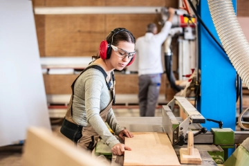 Eine Schreinerin mit Gehörschutz und Schutzbrille arbeitet in einer Werkstatt mit Holz.